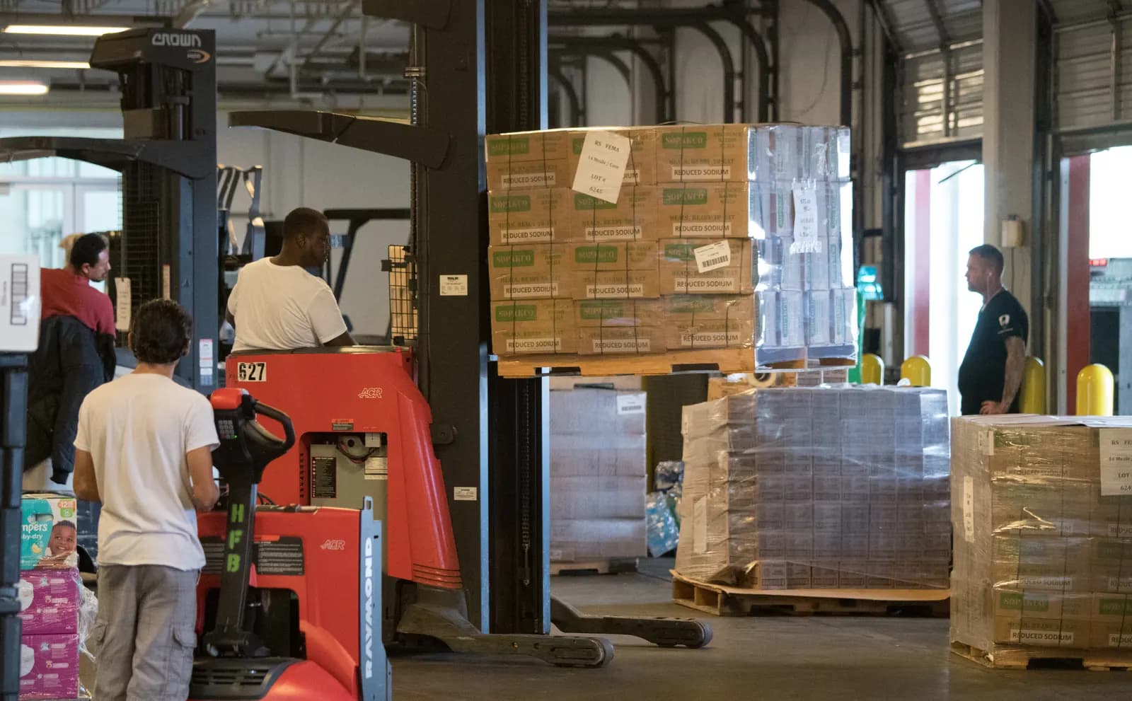 Pallets of boxed goods stacked on racks in a large distribution warehouse with a forklift in the aisle