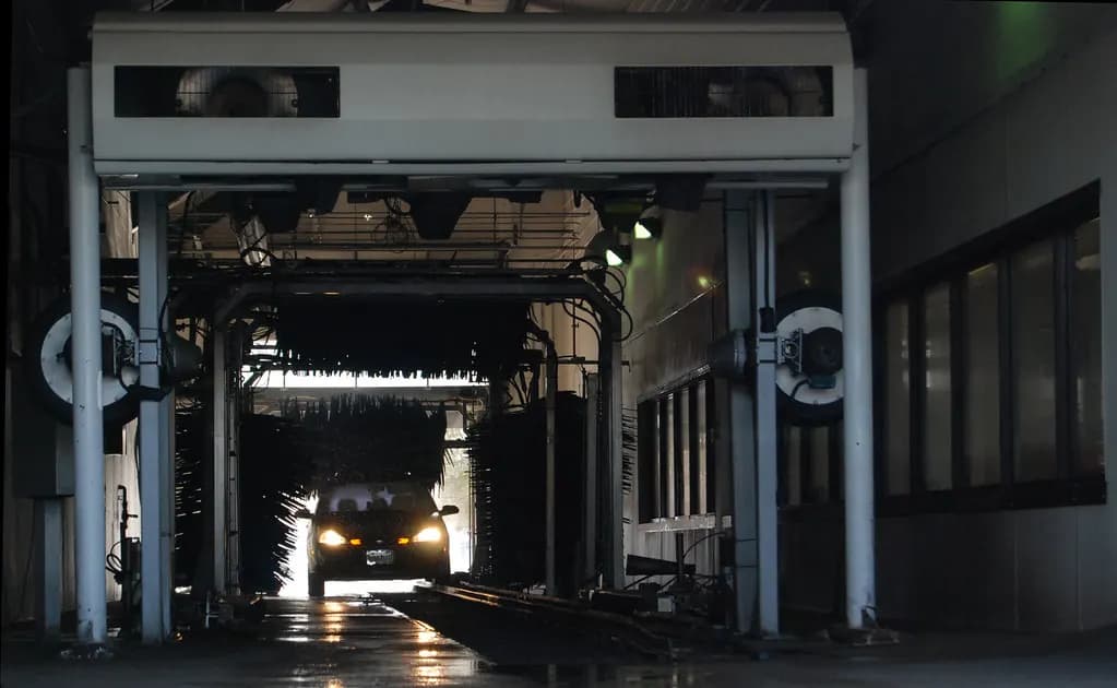 A car with headlights on moving through a conveyor car wash tunnel lined with brushes and dryers