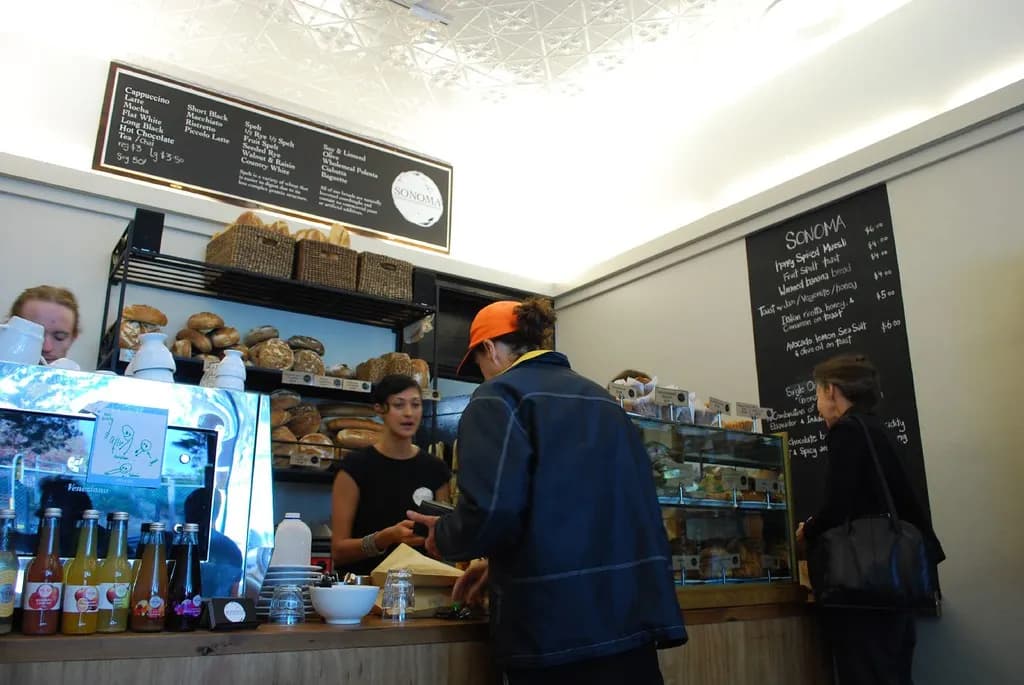 Customers ordering at a small cafe counter with a chalkboard drinks menu, a bread rack and a pastry case