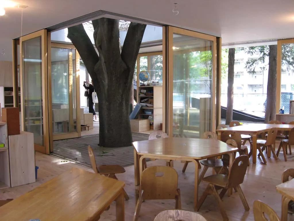 A bright early-years classroom with child-sized wooden tables and chairs, low shelves and glass walls