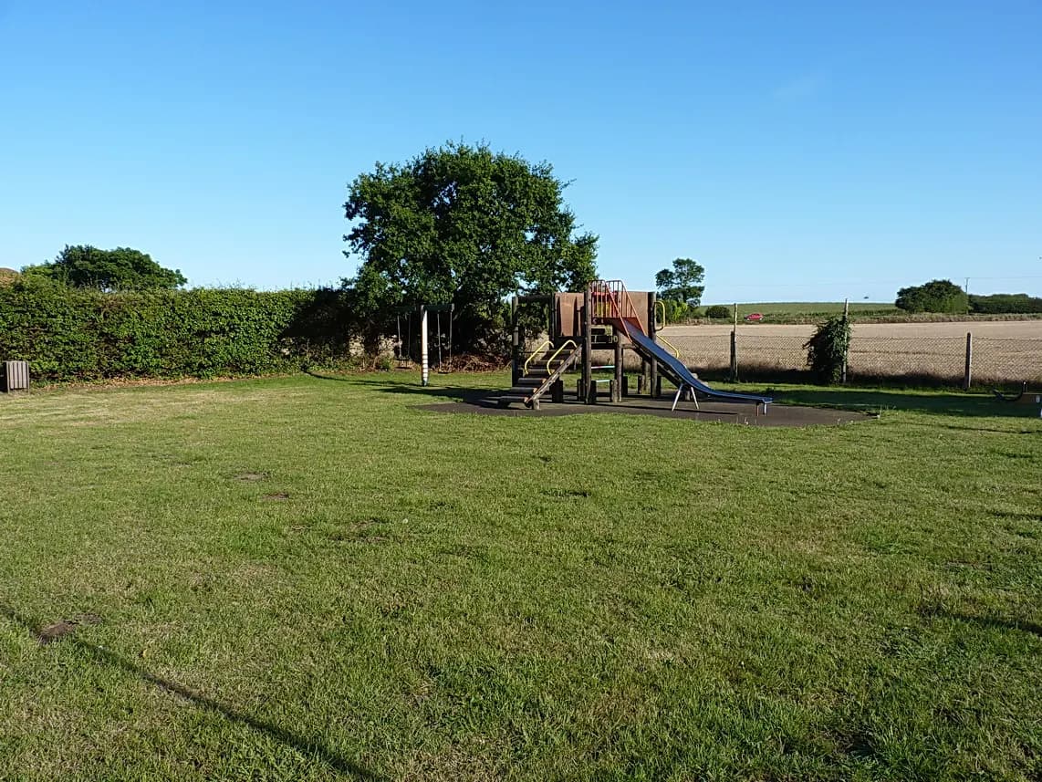 A grassy outdoor play area with a small climbing frame and slide beside a hedge