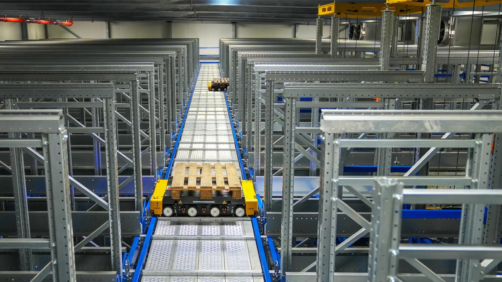 Pallets moving along a conveyor between rows of steel racking in an automated warehouse