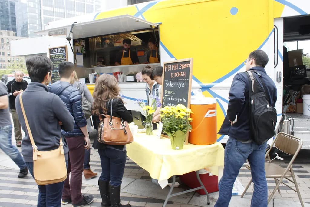 Customers lining up to order at a yellow food truck in Boston with a chalkboard menu and staff cooking inside