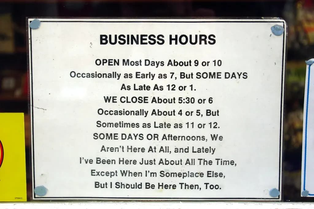 A handwritten-style business hours sign taped inside a shop window listing vague, changing opening times