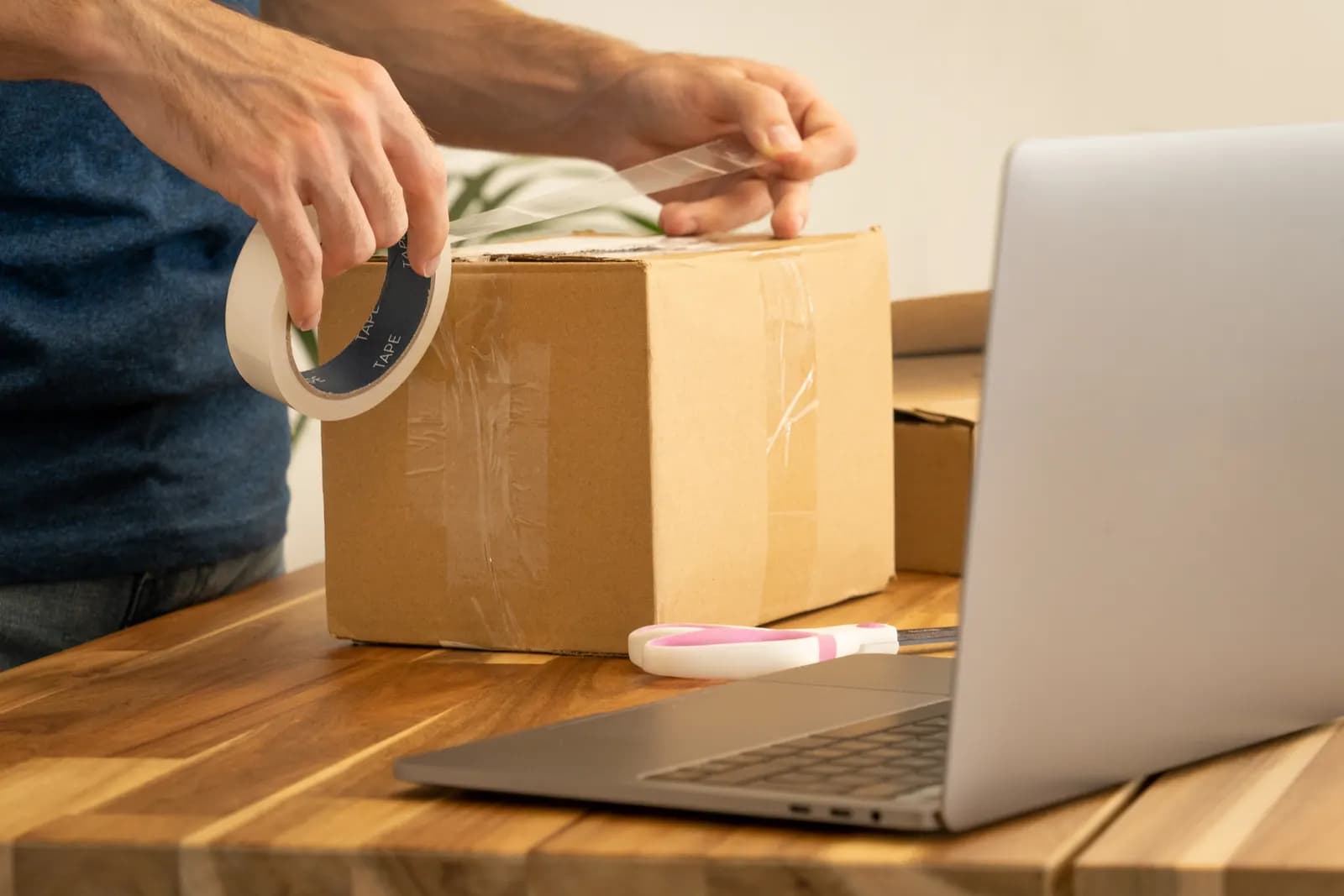 Hands taping a cardboard parcel next to a laptop open on a shipping booking page