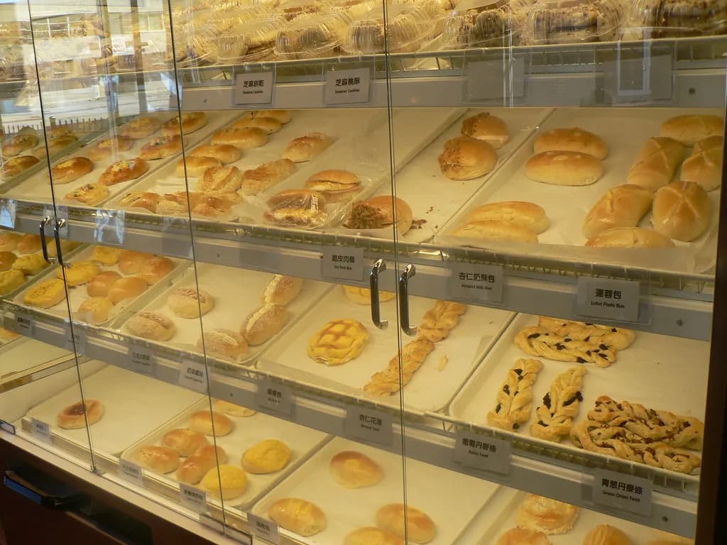 A glass bakery display case with shelves of buns, rolls and pastries on trays