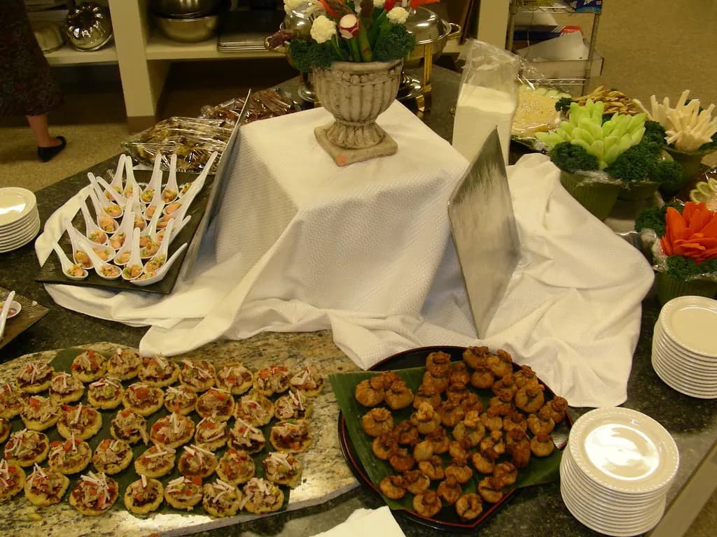 A catered appetizer station with trays of small bites, stacked plates and a white tablecloth