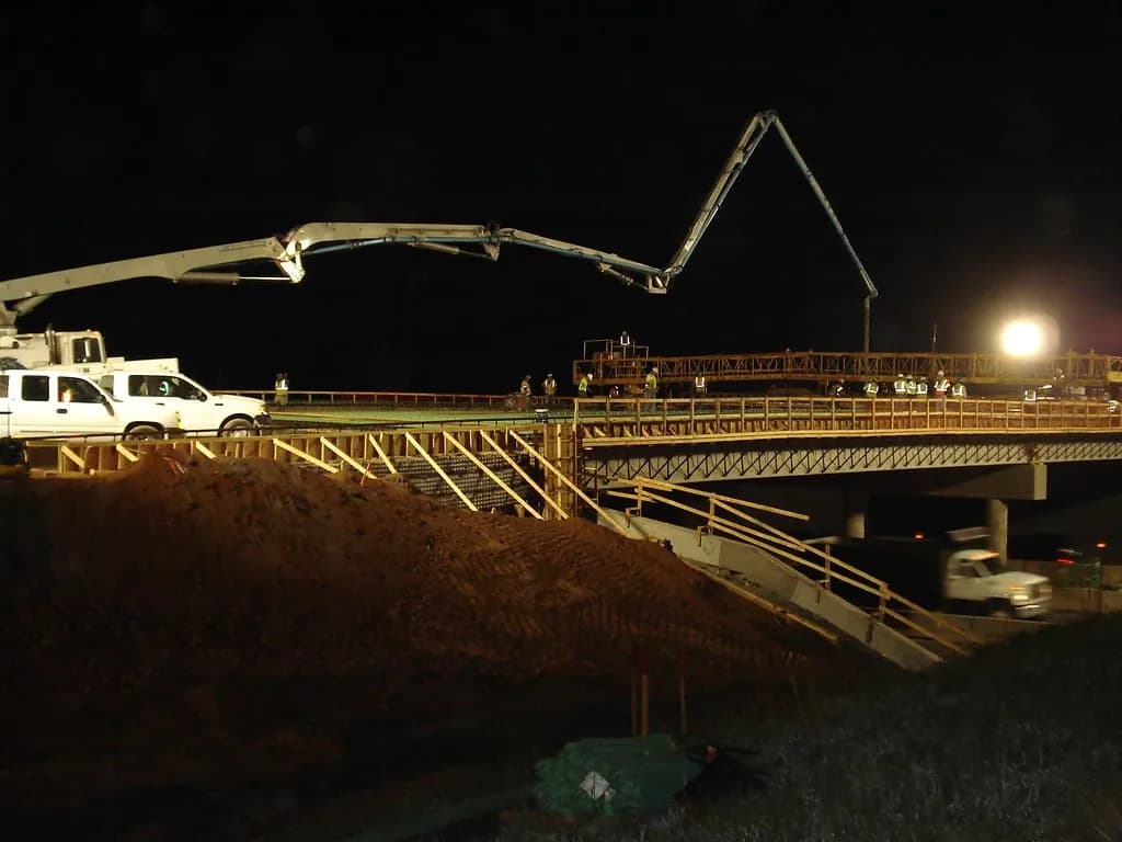 A concrete pump truck with its boom extended placing concrete on a bridge deck at night under work lights