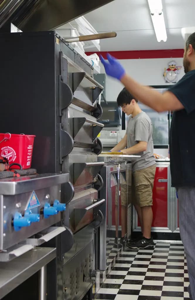 Inside a food truck kitchen: stacked pizza ovens under a stainless hood, with gloved staff preparing food along a narrow aisle