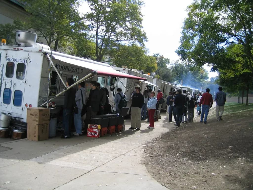 A row of food trucks parked along a tree-lined street with customers queuing at the service windows
