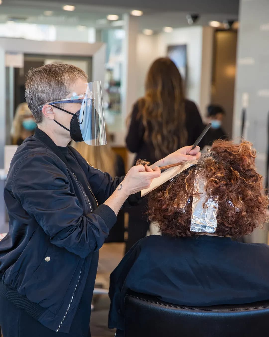 A colorist wearing a face shield applies foil highlights to a client's curly hair at a salon station