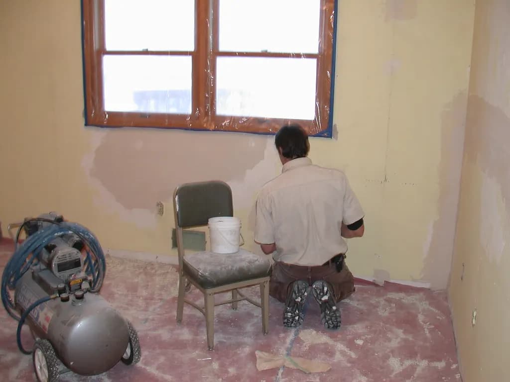 A repair worker patching and sanding a drywall wall in an empty room with an air compressor and a bucket of compound