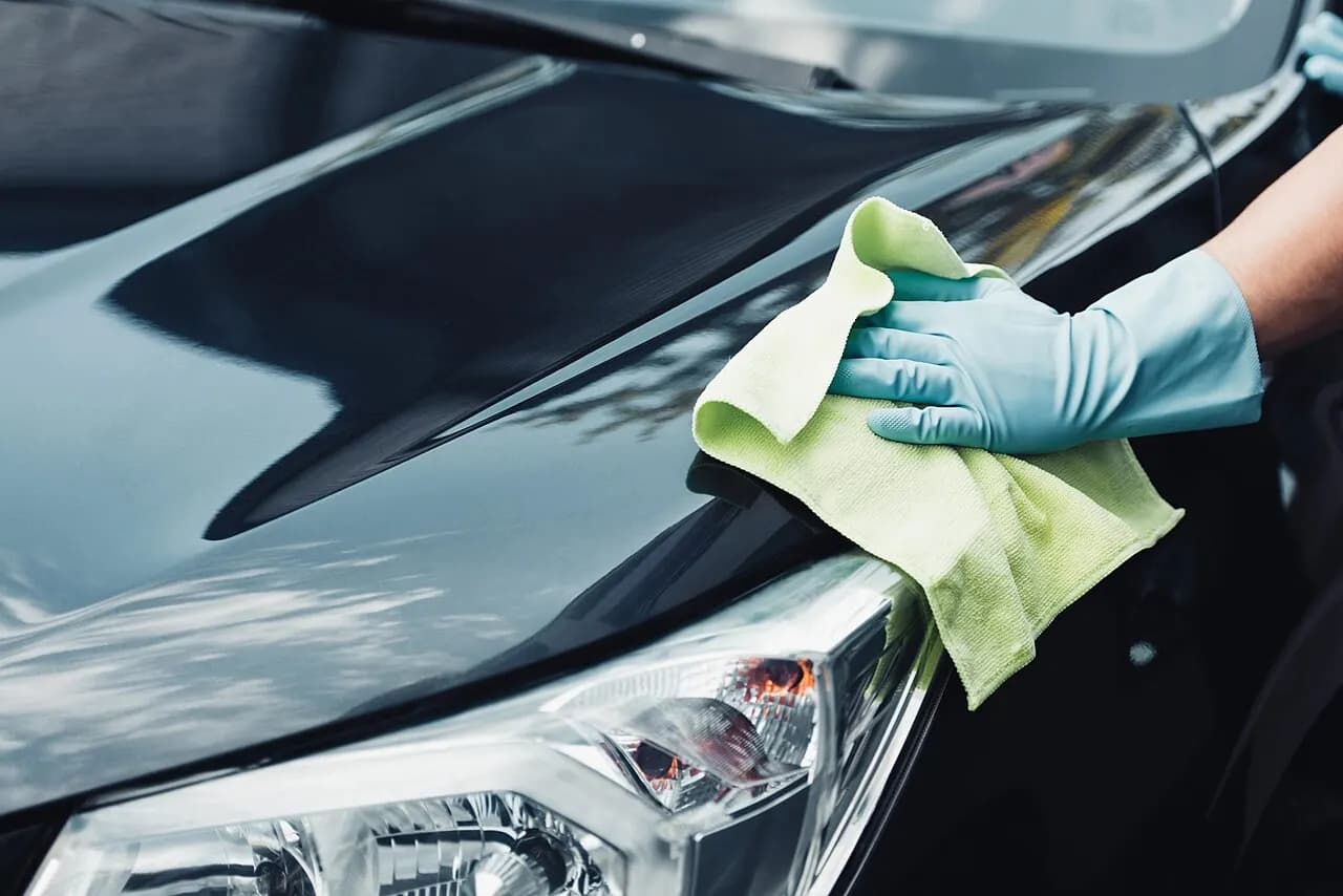 A gloved hand wiping a dark car hood with a yellow microfiber towel next to the headlight
