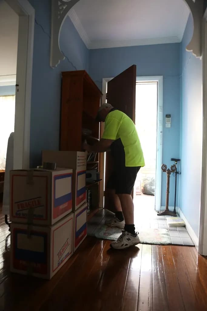 A mover packing books from a wooden bookcase into stacked fragile-marked moving boxes in a hallway