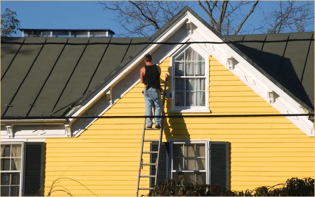 A painter on a ladder painting the yellow clapboard gable of an older wood-frame house