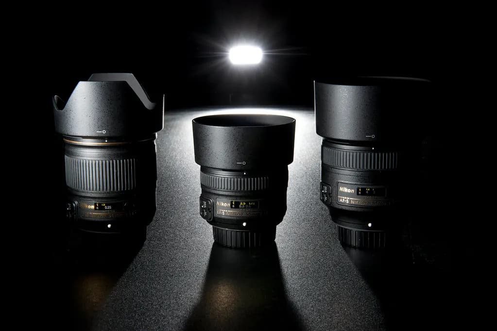 Three prime camera lenses with lens hoods lined up on a dark surface under a single light
