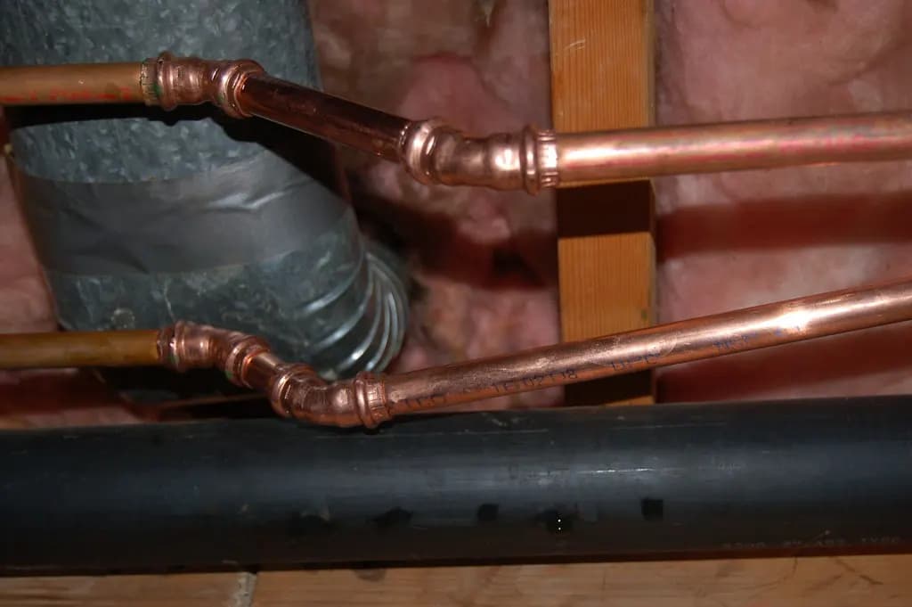 New copper water supply pipes with soldered elbow bends running past a black drain pipe between insulated wall framing