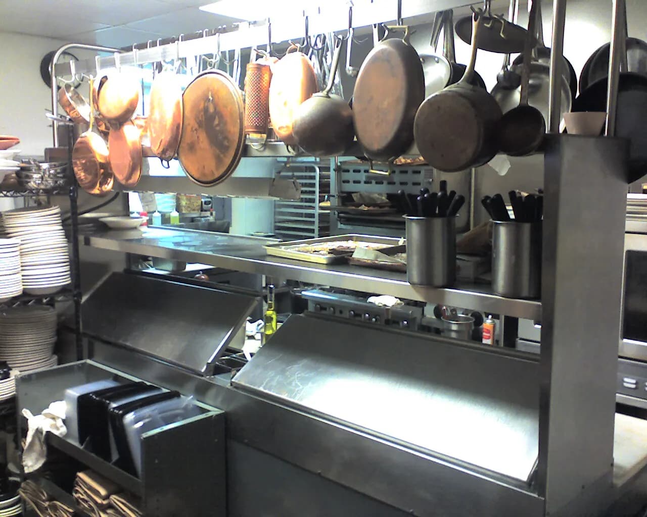 Copper and steel pans hanging above the stainless steel pass in a restaurant kitchen, with stacks of plates at the side