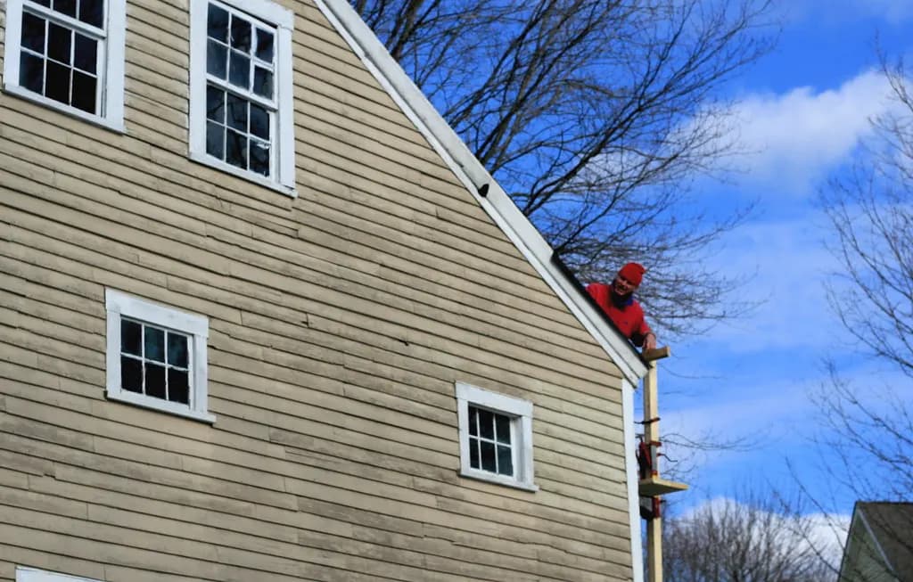 A roofer working at the edge of a steep gable roof on an older clapboard house, with a ladder below