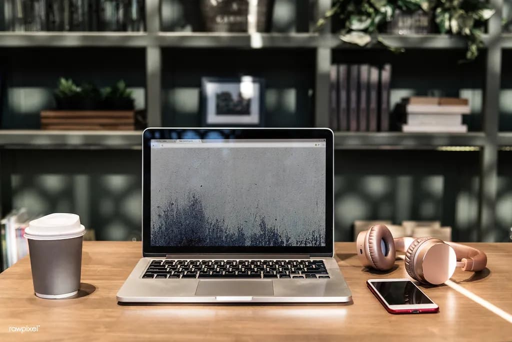 A laptop, coffee cup, phone and headphones on a wooden desk in a coworking space