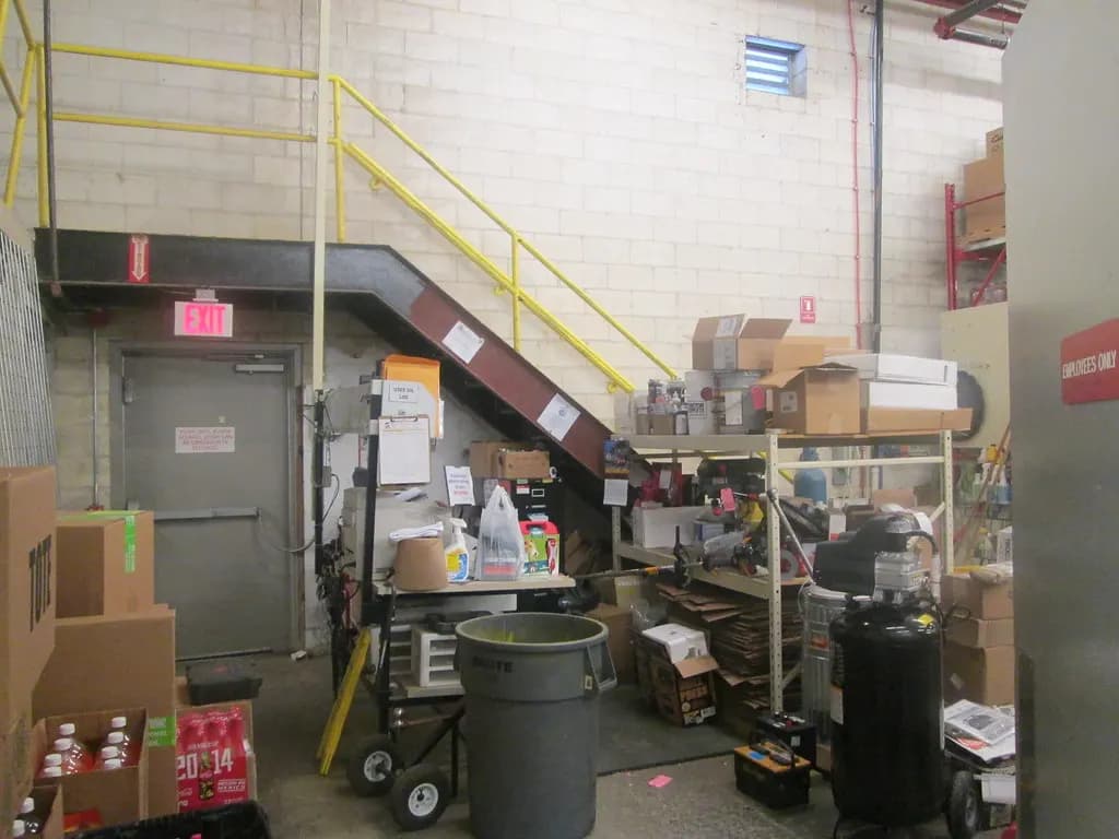Back stockroom of a store with cartons stacked on shelves, a hand truck, bins and supplies near a stairway