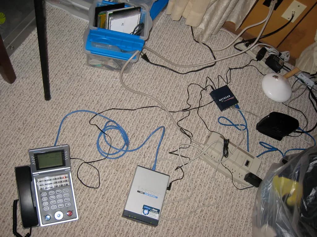 A small office's router, network switch, modem, desk phone and tangled cables lying on a carpeted floor