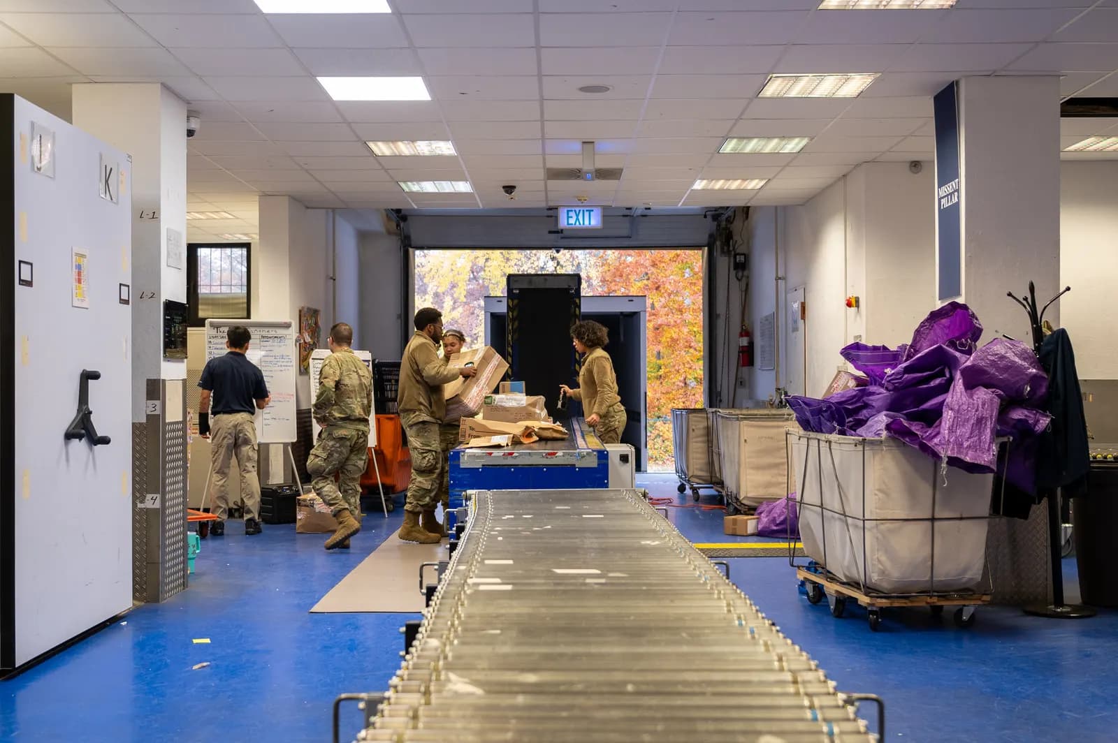 Postal clerks unloading parcels onto a roller conveyor in a mail room, with bins of packages on the right