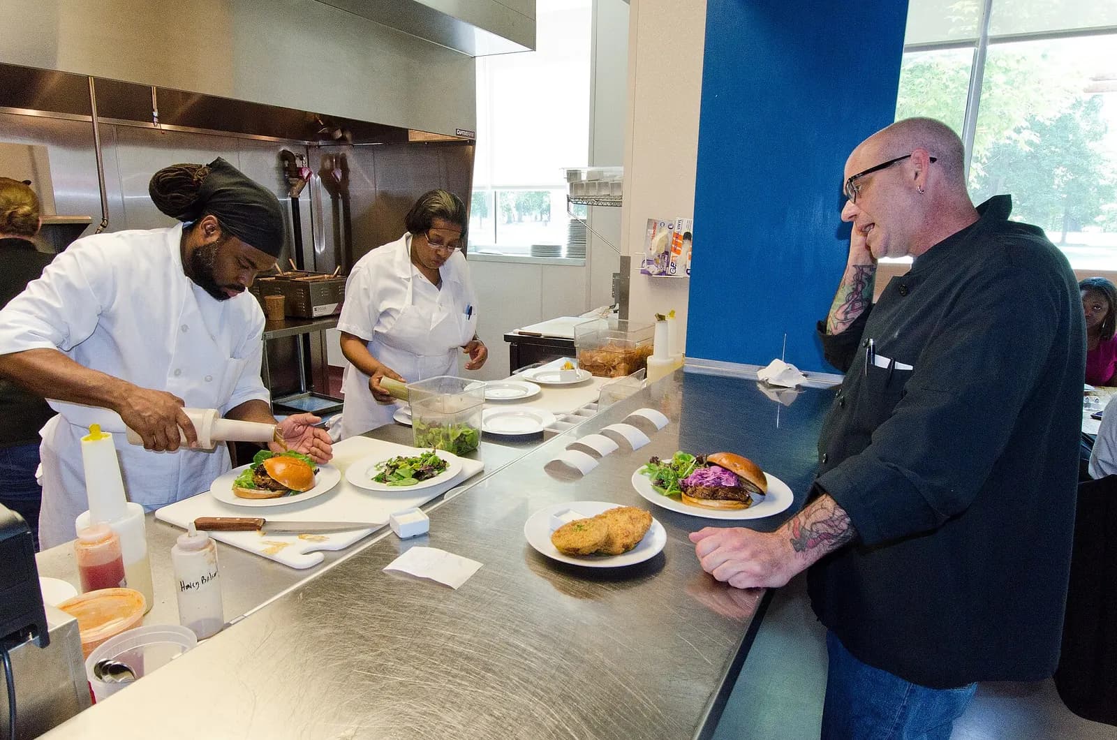 Two line cooks in white jackets plating burgers and salads at a restaurant pass while a chef reads orders