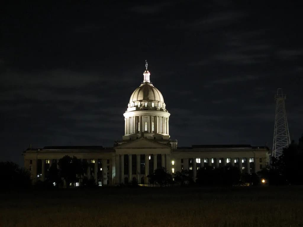 The Oklahoma State Capitol dome lit at night beside an oil derrick on the capitol grounds