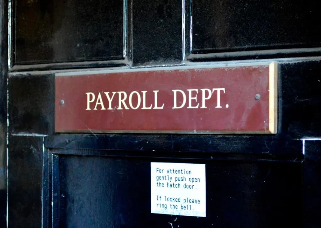 Brass-edged sign reading Payroll Dept. on a black wooden door with a note to push the hatch door