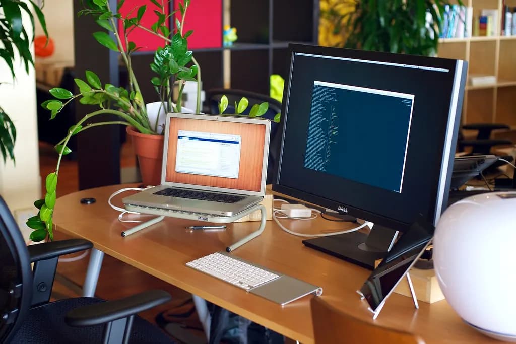 A laptop and a large monitor showing code on a wooden office desk with plants behind