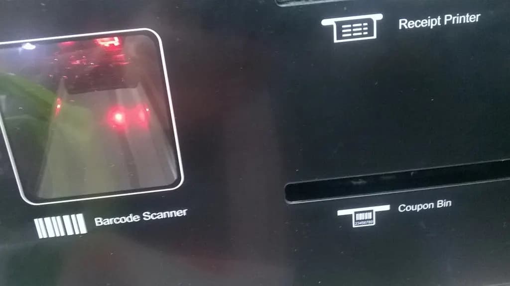 Self-checkout panel at a store with labelled barcode scanner, receipt printer slot and coupon bin