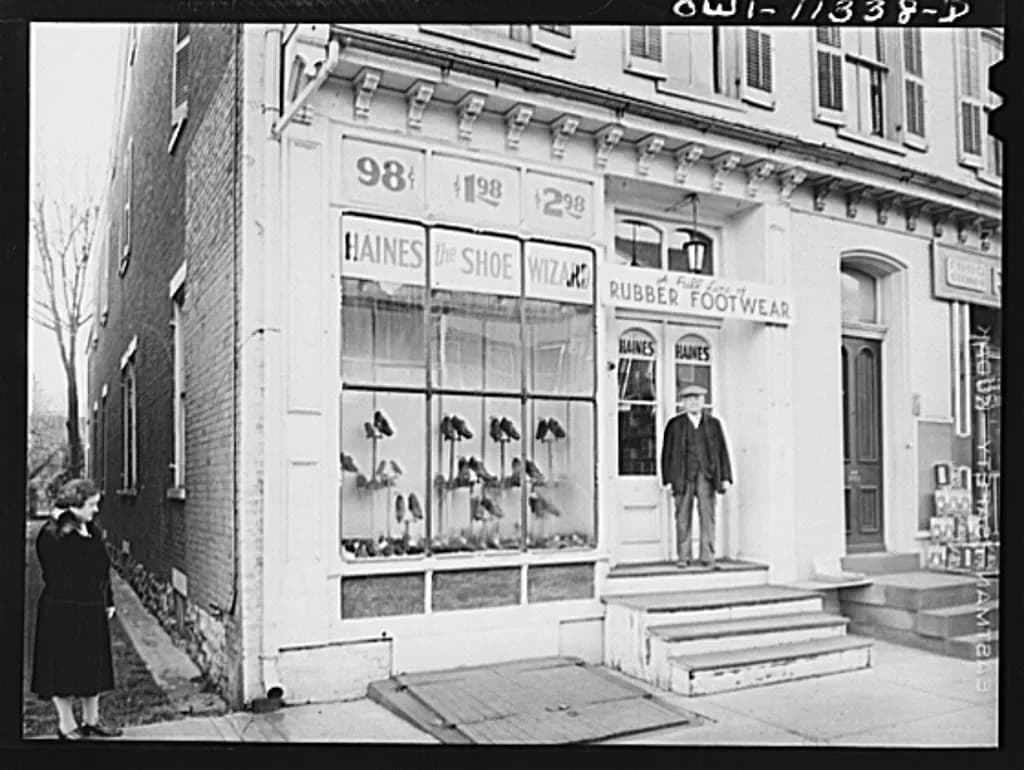 Black-and-white photo of a small shoe store on a town main street with its owner standing in the doorway