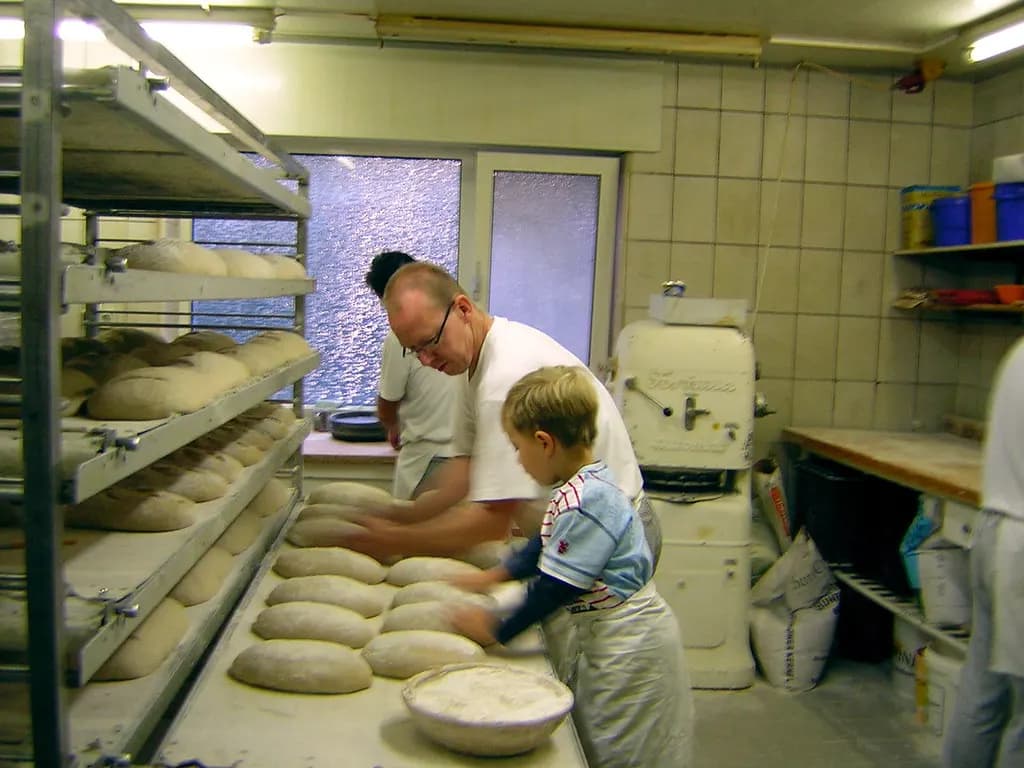 Baker and his young son shaping bread loaves on a floured table beside racks of proofing dough in a small family bakery