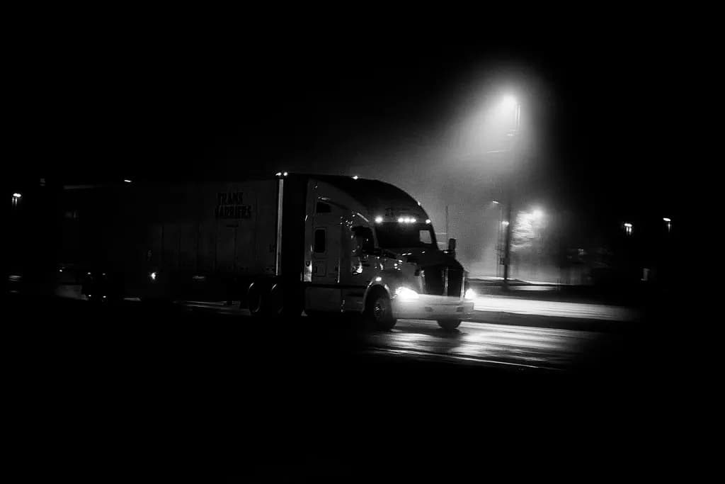 A tractor-trailer driving through rain and fog at night under street lights