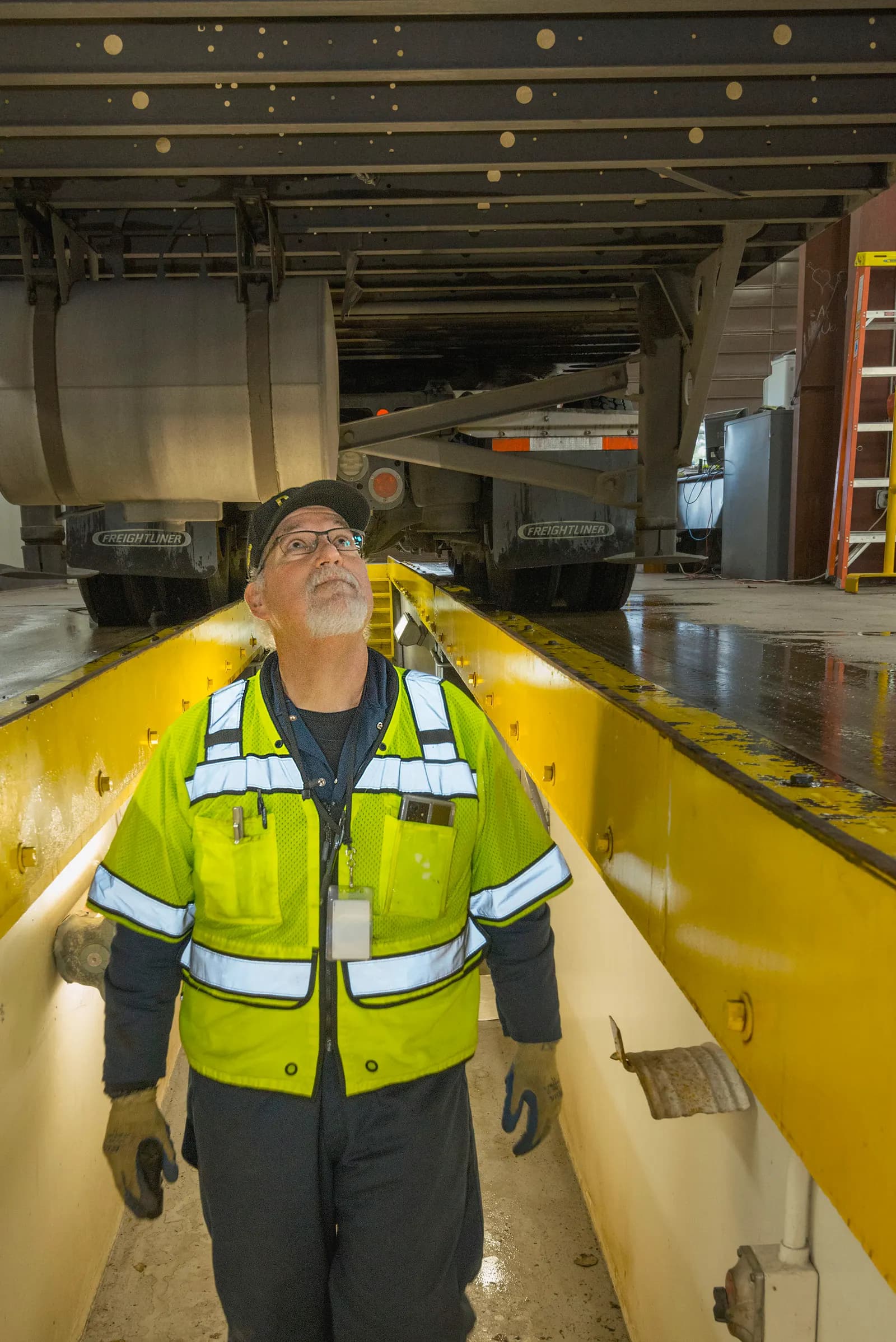 A state inspector in a high-visibility vest checking the underside of a semi-trailer from an inspection pit