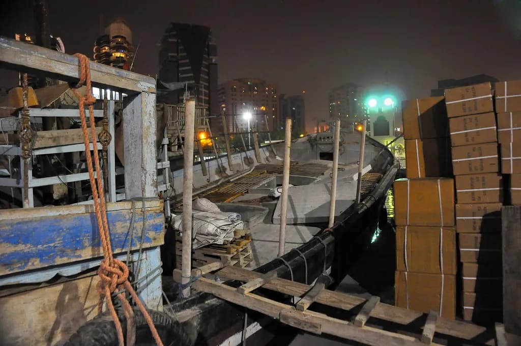A wooden trading dhow at night on Dubai Creek beside stacked cartons of cargo
