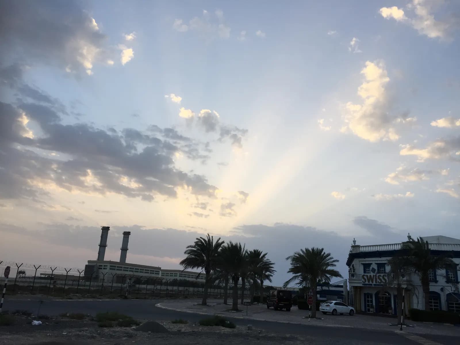 Road inside Hamriyah Free Zone in Sharjah with palm trees, an office building and industrial chimneys under a cloudy sky