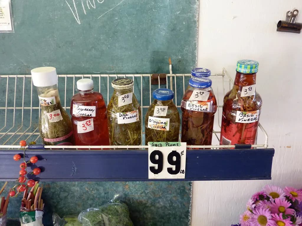 Bottles of herb and fruit vinegars with handwritten labels and price tags on a shelf at the Kelowna farmers' market