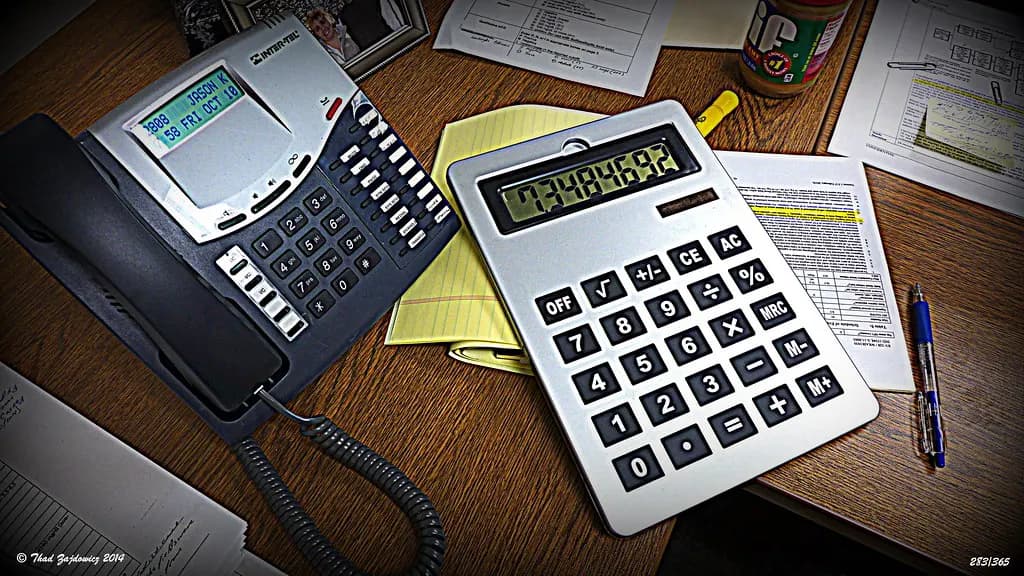A large desk calculator beside an office phone, notepads and printed figures