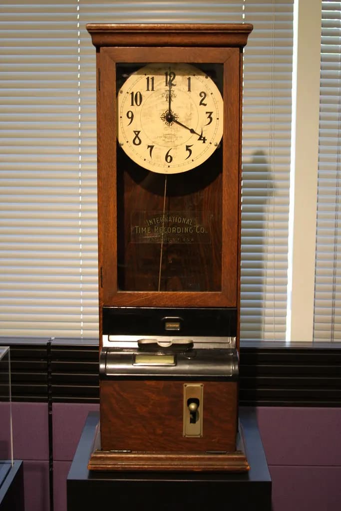 An antique wooden time clock with a round dial and a card slot, displayed against window blinds