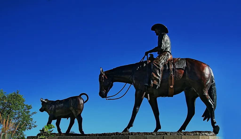 Bronze sculpture of a cowboy on horseback driving a calf, against a clear blue Calgary sky