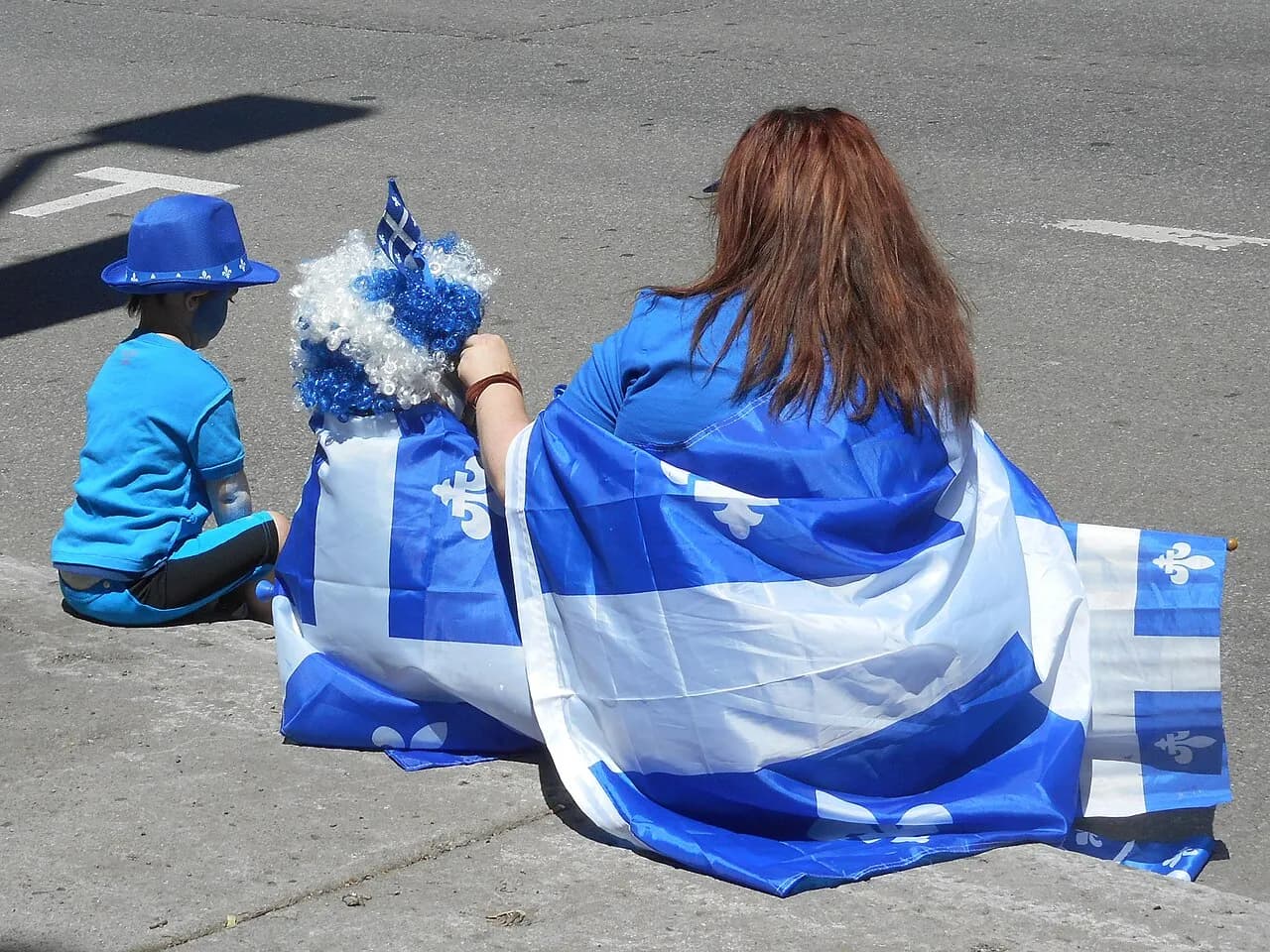 Two spectators wrapped in Quebec flags sit at the curb on rue Saint-Denis in Montreal during Fête nationale celebrations