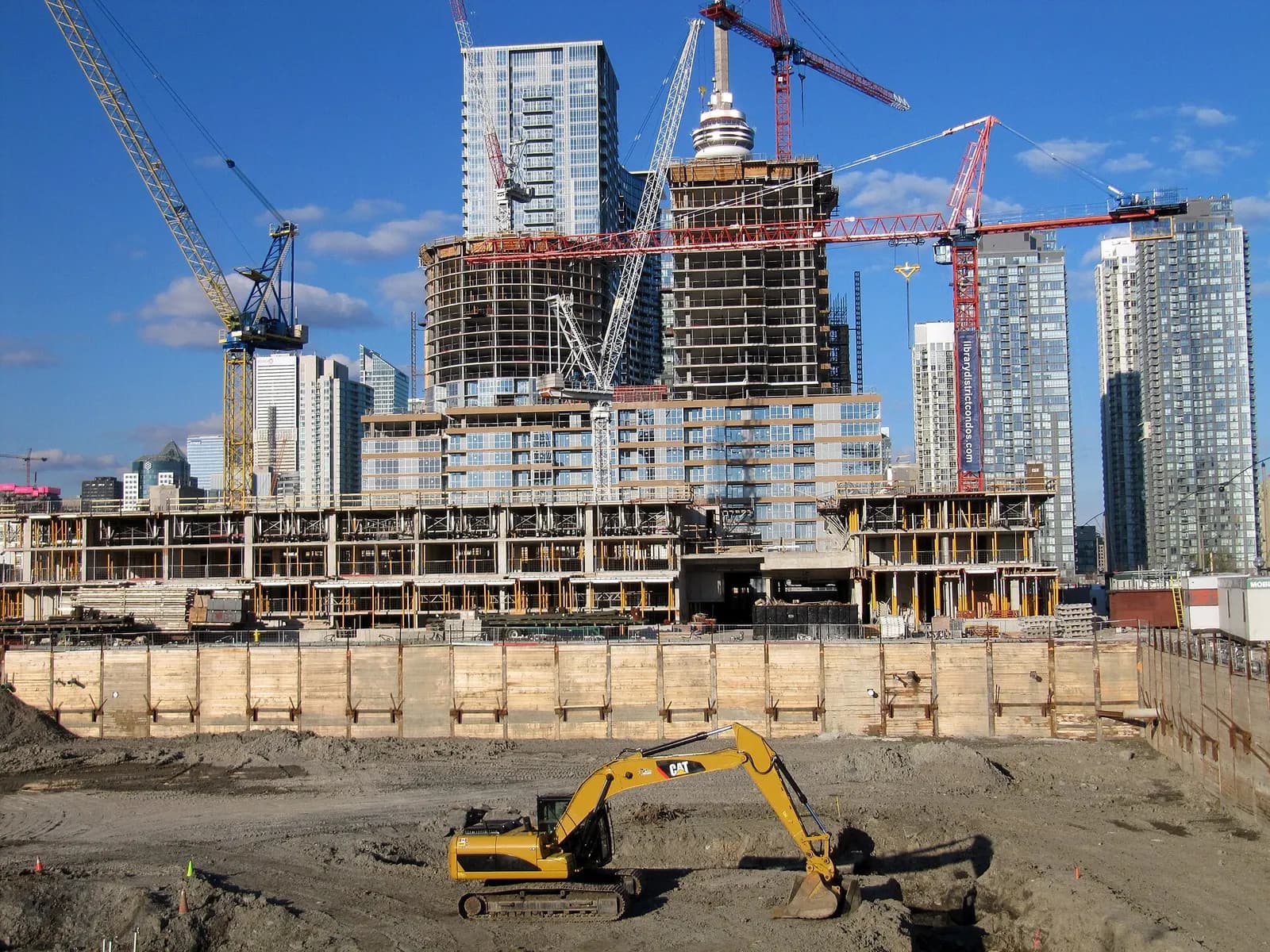 Tower cranes over condominium towers under construction in downtown Toronto, with an excavator in the foreground