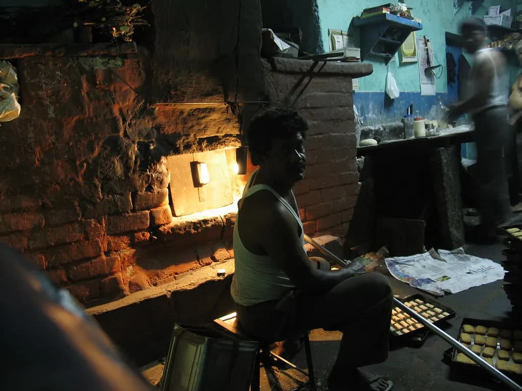A baker sitting by a brick oven in a small Indian bakery at night, with trays of buns cooling beside him