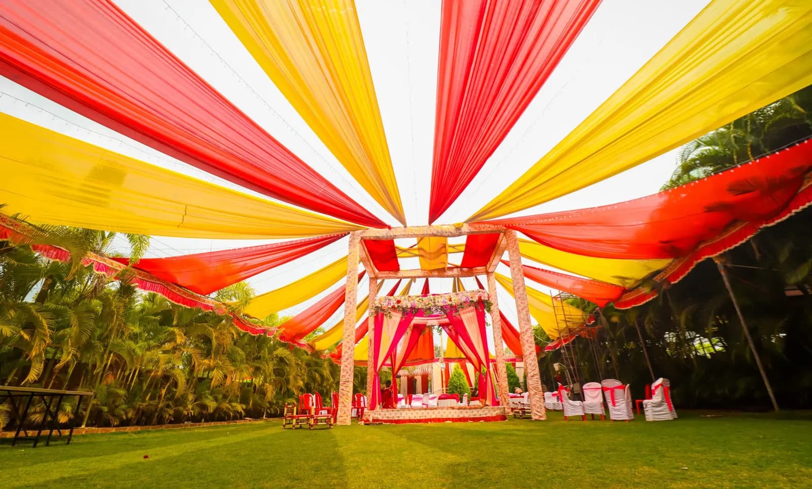 Outdoor wedding mandap on a lawn under red and yellow fabric canopies, with chairs set out for guests
