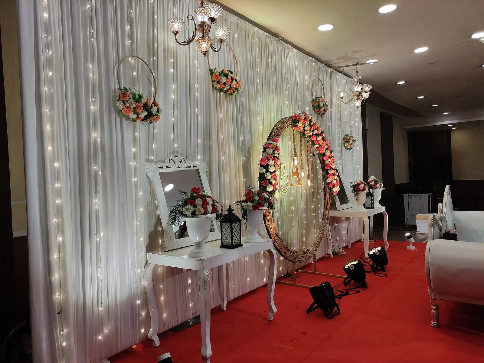 Wedding reception stage in Kochi with white drapes and fairy lights, floral hoops, white side tables and floor uplights on a red carpet