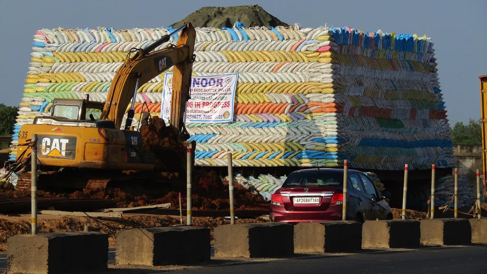 A tall wall of stacked cement bags beside an excavator at a road construction site in India