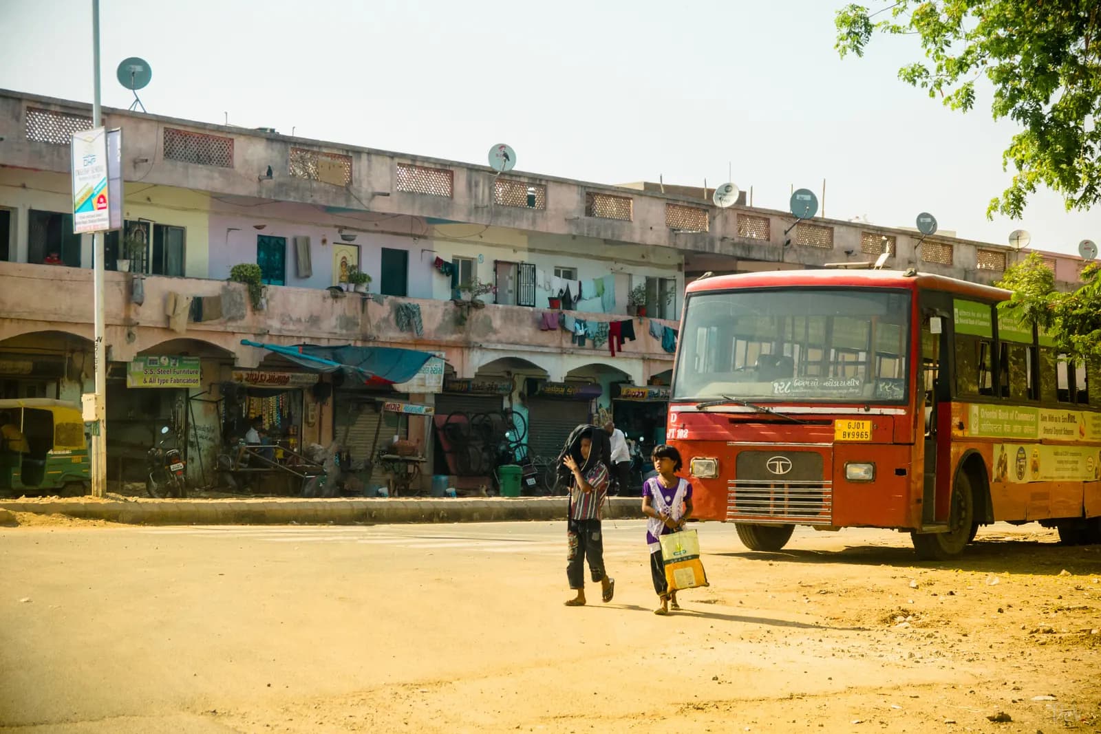 Two children carrying bags walk across a road in front of a red state transport bus in Gujarat