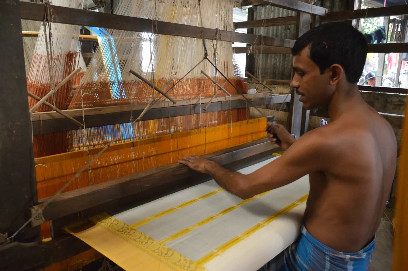 A weaver working a wooden handloom to weave a white tant saree with a yellow border in Phulia, Nadia, West Bengal